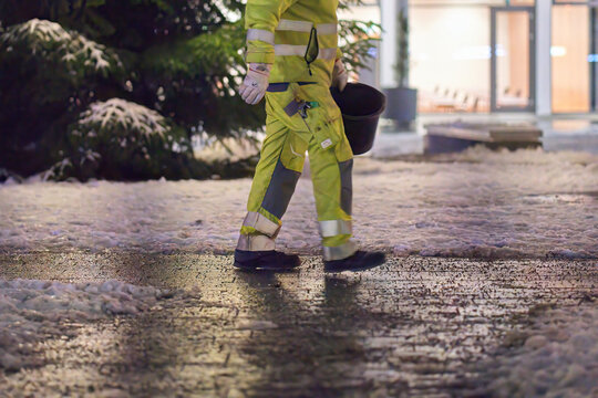Winter service worker spreads defrost salt on the slippery and icy (Blitzeis, Glatteis) city sidewalk. Man with yellow uniform on frozen road. Christmas time in early winter morning. Germany.