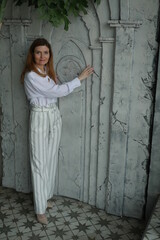 Stylish woman in white shirt and trousers posing standing next to textured wall