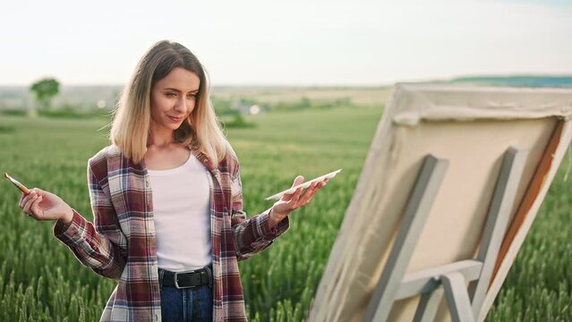 Side view of female artist drawing, painting on easel, enjoying, admiring. Blonde, cheerful woman wearing plaid shirt and jeans, working. Concept of art and modern lifestyle.