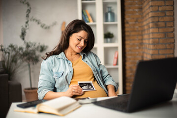 Beautiful young pregnant woman holding ultrasound pictures of her baby. Businesswoman having video call..