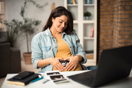 Beautiful young pregnant woman holding ultrasound pictures of her baby. Businesswoman having video call..