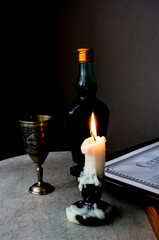 A burning candle on the table in front of a glass and a bottle. Soft focus