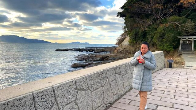 A woman walking while holding and drinking a cup of hot coffee by the sea during sunset and cold weather