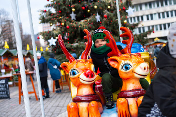 Smiling cheerful kid boy rides a reindeer carousel in the background of a Christmas tree - 555142970