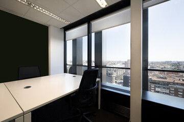 Several white empty corporate work desks with swivel armchairs and large windows overlooking the city
