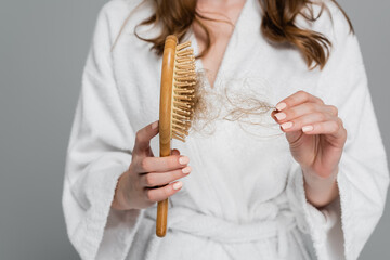 cropped view of young woman holding wooden hair brush and pulling damaged hair isolated on grey