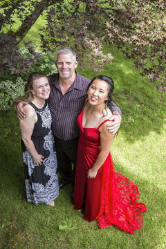 Portrait Of A High School Graduate Wearing A Red Formal Dress Standing With Her Parents In A Yard; Chilliwack, British Columbia, Canada