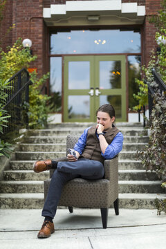 A young man sits in an armchair outdoors using his smart phone; Bothell, Washington, United States of America