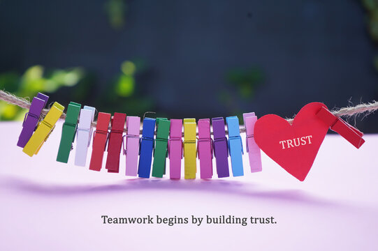 Inspirational and motivational quote - Teamwork begins by building trust. With text TRUST on red heart and colorful wooden clips on rope on dark and light pink purple background. Business concept.