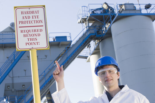 Lab worker pointing at a warning sign saying 'Hardhat and Eye Protection Required Beyond This Point' at an asphalt plant