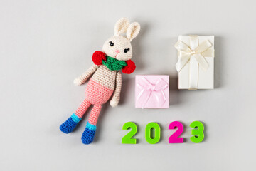 Knitted children's toy bunny or rabbit with pink and beige gift boxes and numbers 2023 on a gray background. The symbol of 2023.