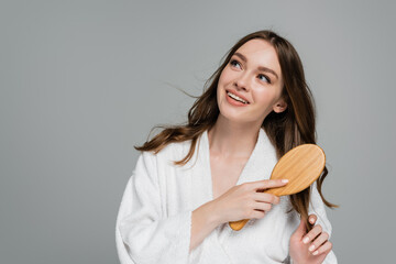 pleased young woman holding wooden hair brush while brushing shiny hair isolated on grey