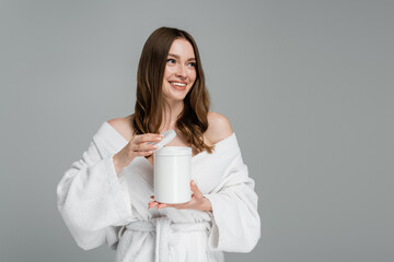 joyful young woman with shiny hair holding container with mask isolated on grey