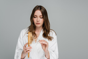 stressed young woman holding wooden hair brush and pulling damaged hair isolated on grey
