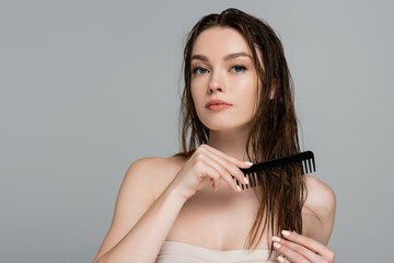 pretty young woman with bare shoulders brushing wet hair with comb isolated on grey
