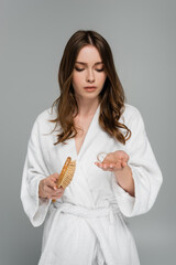 sad young woman in bathrobe holding hair brush and damaged hair isolated on grey