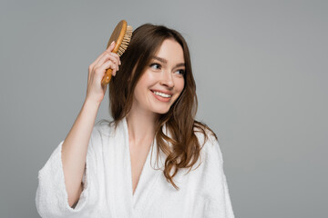 cheerful young woman brushing shiny and healthy hair while looking away isolated on grey