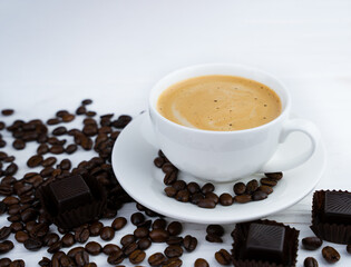 Cup of fresh coffee, chocolate candies and scattered coffee beans on a white wooden background. Coffee day. Close-up. Copy space. Selective focus.