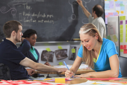 Engineering instructor working on flowchart on blackboard while students working on project, one man with spinal cord injury and other one with Aspergers