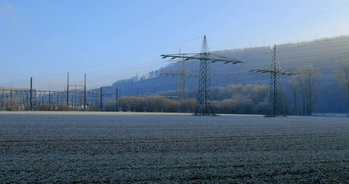 Würgassen, Germany - 12/17/2022: High-voltage pylons with overhead lines and switchgear with blue sky in winter with a field and ice