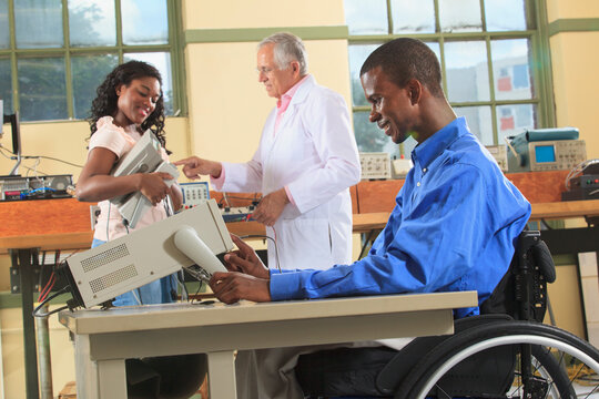 Professor and engineering students in an electronics classroom, one in a wheelchair preparing oscilloscope for the lab