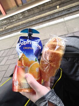 White womans hand holding an orange capri-sun drink pouch and all butter croissant in front of a holdall bag and rucksack on a train platform.
