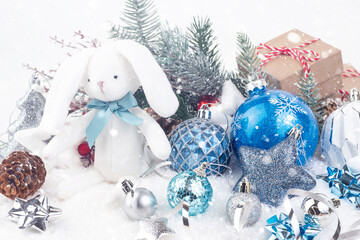 Fototapeta na wymiar White rabbit with a blue bow around its neck and various Christmas decorations on a white background. In the foreground are cones and Christmas decorations. The concept of New Year and Christmas. 