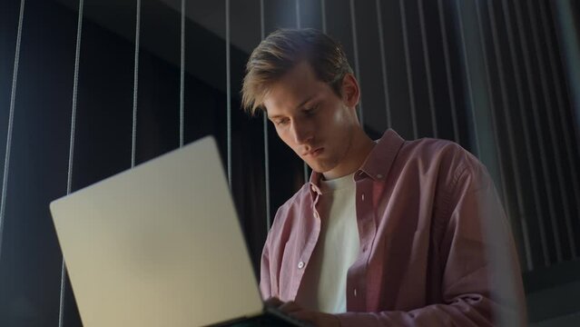 Low-angle view of focused freelancer male working at laptop in home office. Successful young designer edit text or images in professional program. Confident businessman working distantly in dark room.