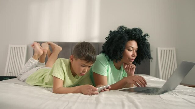 Young black African American university student learning online using laptop computer with her son laying near in bedroom. Woman taking notes, watching webinar, virtual education remote class studying