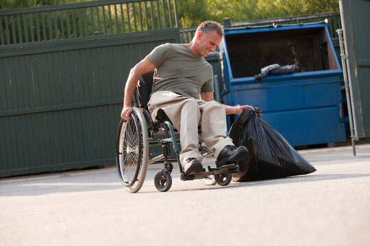 Man in wheelchair with Spinal Cord Injury trying to move heavy trash bag to dumpster