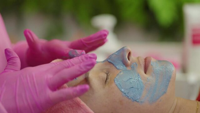 Close-up shot of a woman enjoying facial treatment with clay mask at beauty shop. Cosmetology and spa

