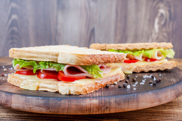 Two sandwiches made with grilled bread filled with cheese, slice of ham, lettuce and tomatoes served on board on wooden background.