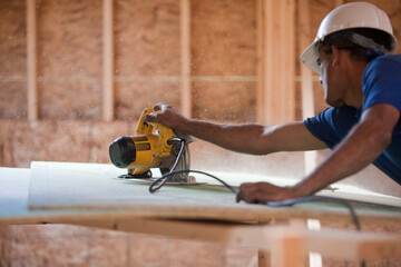 Hispanic carpenter using circular saw on roofing sheathing at a house under construction