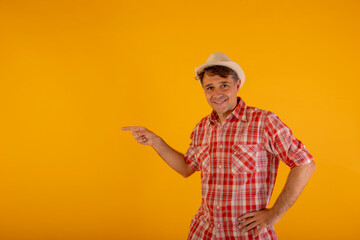 Man in hat and colorful checkered shirt pointing to the side with his hands on an orange background