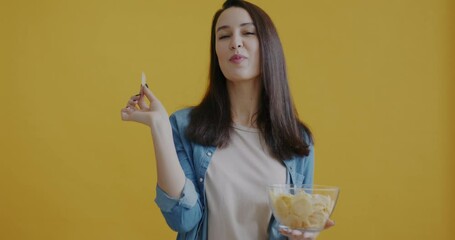 Portrait of joyful young woman eating potato chips with pleasure smiling on yellow color background. Nutrition and delicious snack concept.