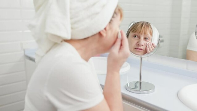 Gorgeous mid age older adult 50 years old blonde woman wears bathrobe in bathroom applying nourishing antiage face skin care cream treatment, looking at mirror doing daily morning beauty routine.