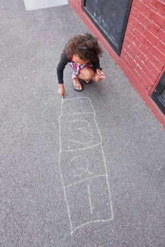 A girl crouches on the sidewalk and draws hopscotch game with chalk