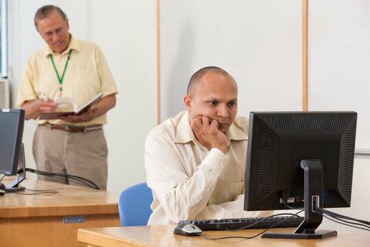 Engineering student studying computer display in a classroom with professor in background