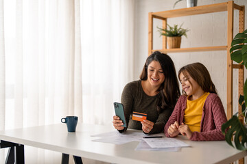 Young mom with school age daughter making purchases in popular internet store from home. Smiling woman holding credit card for online payments.