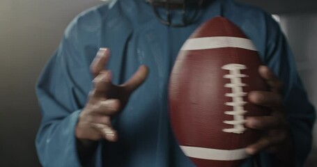 Close-up of African-American man professional rugby player holding ball for American football wearing blue uniform and helmet. Aggressive play with ball in gym. Warm-up concept. - Powered by Adobe
