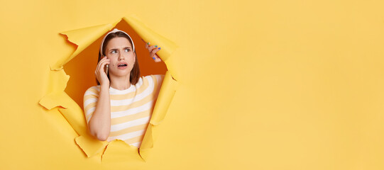 Portrait of shocked woman wearing striped T-shirt, hair band and sunglasses, breaking through paper hole in yellow wall, talking on smart phone, hearing astonishing news. Copy space.