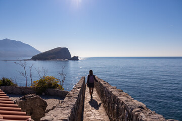 Tourist woman walking on wall of historical citadel of Budva with Scenic view over Saint Nicholas island (Sveti Nikola) and Adriatic Sea, Montenegro, Balkan, Europe. Sunny day at Budvanian Riviera