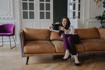 Satisfied young brunette in white shirt and violet pants sitting on leather sofa at office with opened diary, happy, entrepreneur planning agenda. Student woman smiles, relaxing. Business