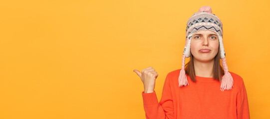 Portrait of sad disappointed woman wearing funny earflap hat and orange casual jumper standing isolated over yellow background, looking at camera with pout lips, pointing at copy space.