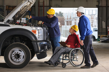 Vehicle mechanic working on a truck and inspectors, including a man in wheelchair, watching him work