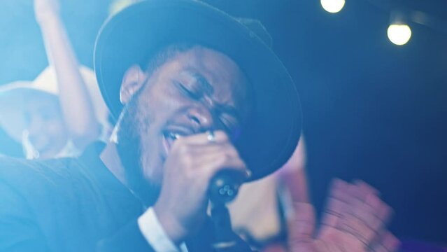 Close-up portrait of energetic happy African singer performing music event singing with microphone vibing. Attractive upbeat man making party for young positive people hanging out on fest.