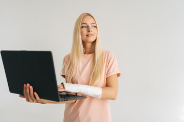 Portrait of smiling injured young woman with broken arm wrapped in plaster bandage holding laptop and working typing, looking thoughtful away, standing on white isolated background.