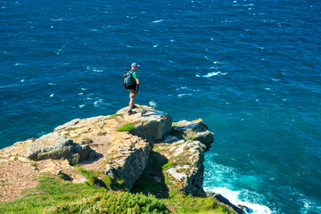 Male hiker on rocky cliff overlooking a sea of blue water; Cornwall County, England