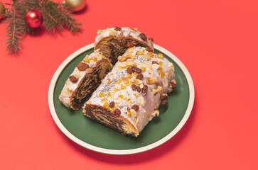 Christmas strudel with poppy seeds,traditional polish festive pie Makowiec on green plate,covered with sugar icing,walnuts,dried fruits on red background with Christmas tree and toy balls. Horizontal