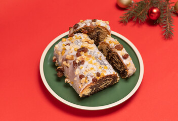 Christmas strudel with poppy seeds,traditional polish festive cake Makowiec on green plate,covered with sugar icing,walnuts,dried fruits on red background with Christmas tree and toy balls. Horizontal